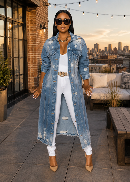 A woman stands on a rooftop patio wearing a long, distressed light-wash blue denim jacket over a white outfit, with sunglasses, layered necklaces, and tan pointed heels. The background shows a city skyline at sunset, a brick wall, and outdoor seating.