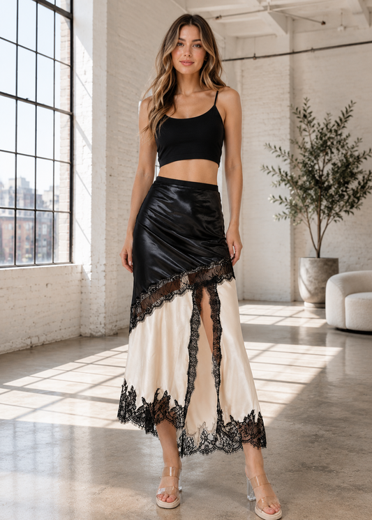 A woman models a two-piece outfit consisting of a black spaghetti-strap crop top and a matching black-and-cream satin skirt with lace trim, standing in a bright loft-style room with large windows.