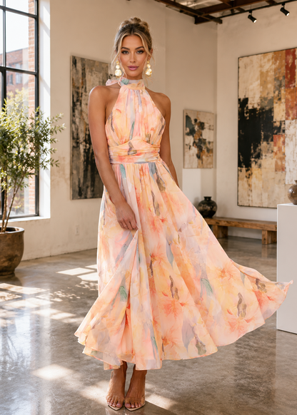 A woman modeling a sleeveless halter-neck maxi dress with a flowing watercolor floral print in pastel peach, orange, pink, and light blue, standing in a bright modern art gallery.