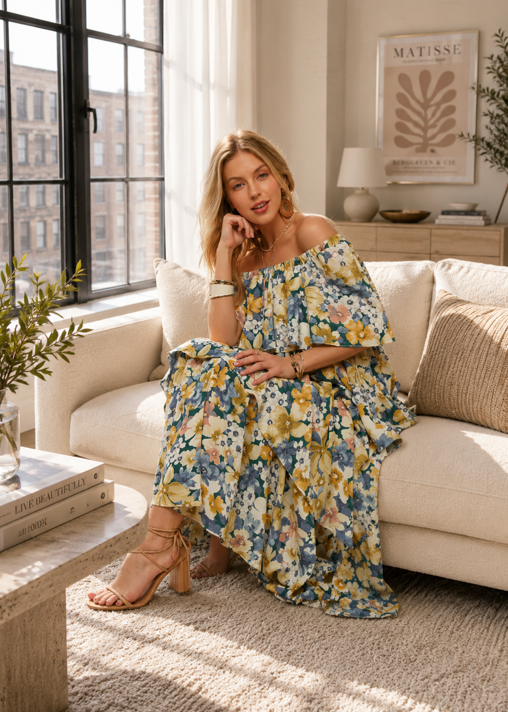 A woman sits on a cream sofa in a bright, modern living room, wearing a blue off-the-shoulder maxi dress with a yellow, white, and pink floral print. She is wearing beige strappy high-heeled sandals.