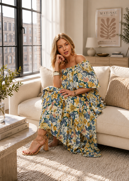 A woman sits on a cream sofa in a bright, modern living room, wearing a blue off-the-shoulder maxi dress with a yellow, white, and pink floral print. She is wearing beige strappy high-heeled sandals.