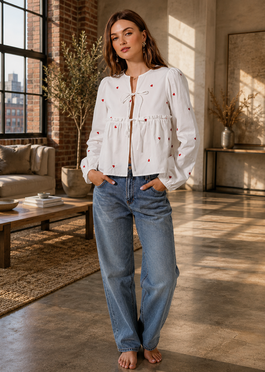 A woman stands barefoot in a sunlit loft room wearing a white long-sleeve blouse with small red embroidered dots and a tie neckline, paired with light blue jeans.