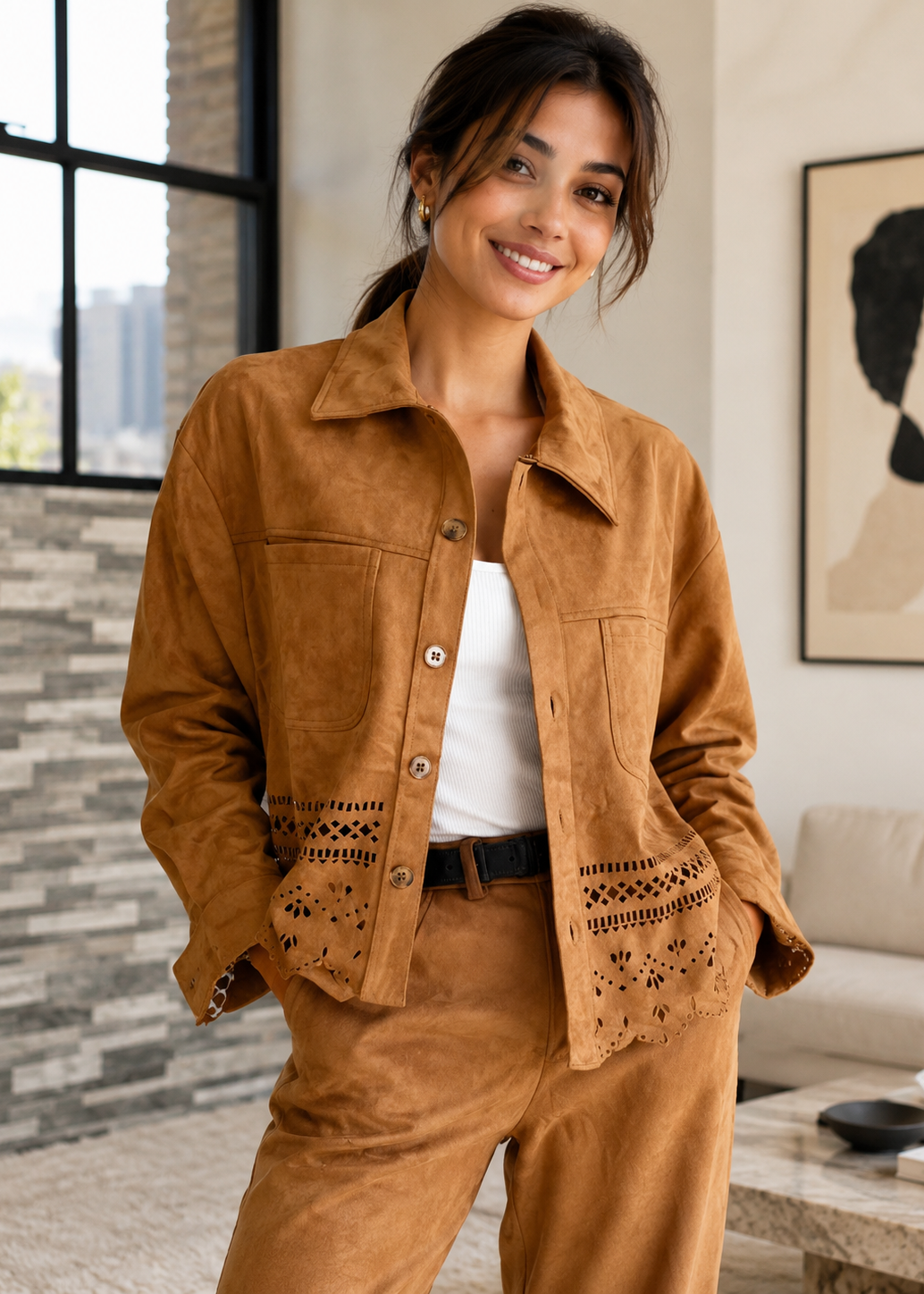 A woman wearing a tan suede-like button-front jacket with decorative cutout detailing on the front pockets, paired with matching tan pants and a white top, standing in a modern indoor room.