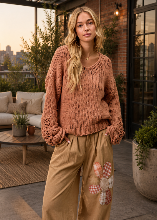 A woman stands on a patio wearing a chunky knit crewneck sweater in a warm terracotta color, paired with tan wide-leg pants featuring a floral patchwork design on the leg.