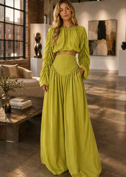 A woman modeling a bright yellow-green two-piece outfit consisting of a long-sleeve crop top and matching high-waisted maxi skirt, standing in a modern loft-style room with brick walls and large windows.