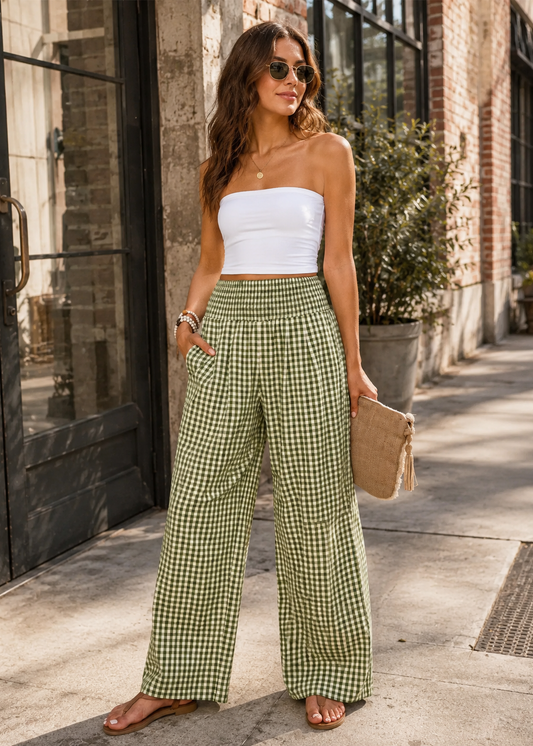 A woman stands on a city sidewalk wearing a white strapless tube top and green-and-white gingham wide-leg pants, holding a beige clutch bag.
