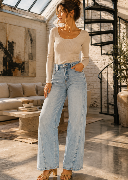 A woman stands in a bright loft-style room wearing a fitted beige long-sleeve top and light-wash wide-leg blue jeans, with tan heeled sandals.