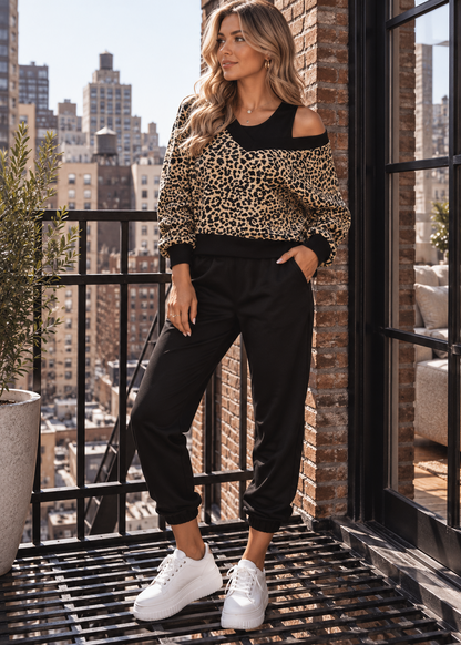 A woman stands on a balcony wearing a leopard-print cold-shoulder top with black trim, black jogger pants, and white sneakers, with a city skyline in the background.