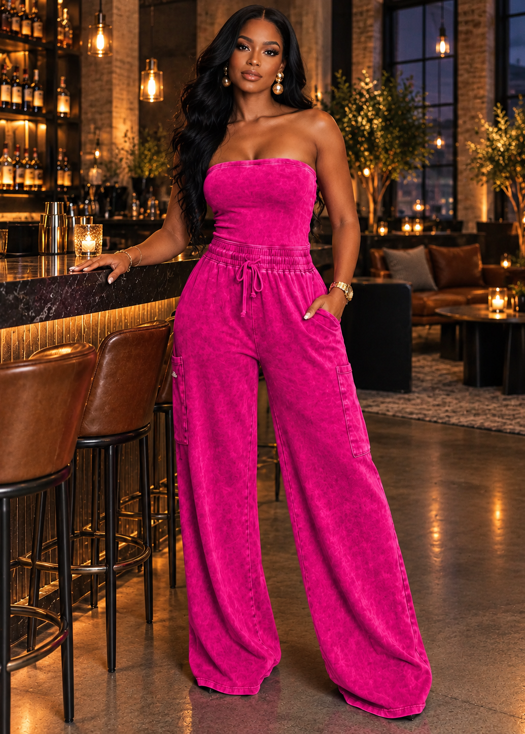 A woman stands in a warmly lit bar or lounge wearing a bright fuchsia pink strapless velvet jumpsuit with wide legs and a drawstring waist. The setting includes bar stools, shelves of bottles, hanging lights, and indoor plants.