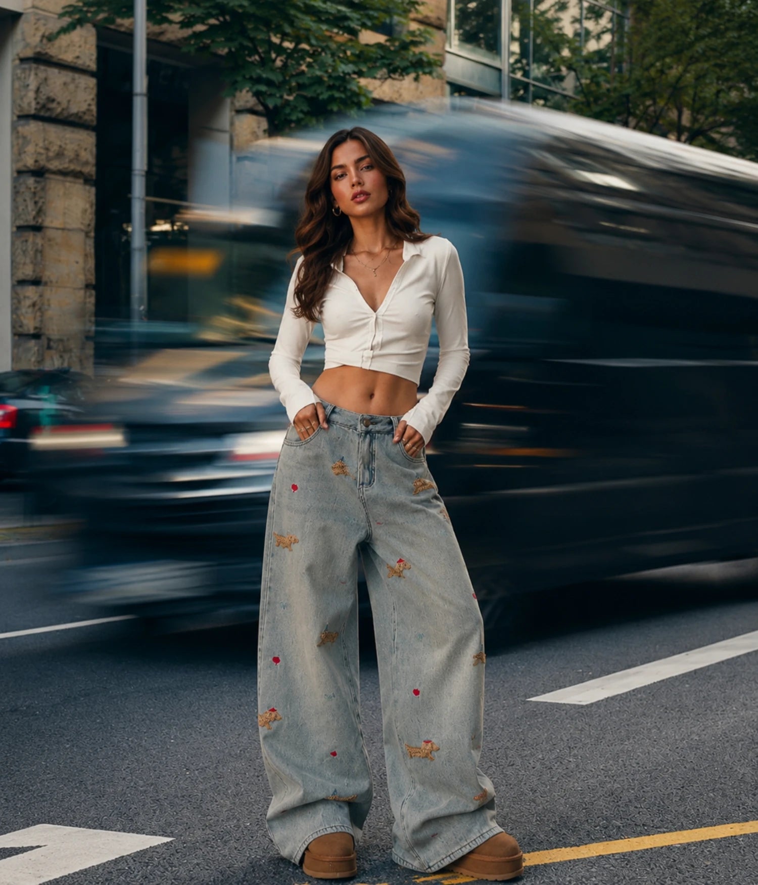 A woman stands on a city street wearing a fitted white long-sleeve crop top and light-wash wide-leg jeans with small embroidered details, posed in front of a blurred passing car.

